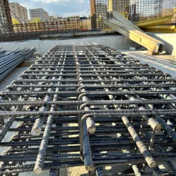 Siatka zbrojeniowa ułożona na betonowej powierzchni, widoczne końcówki prętów i elementy szalunku w tle, w oddali budynki mieszkalne.