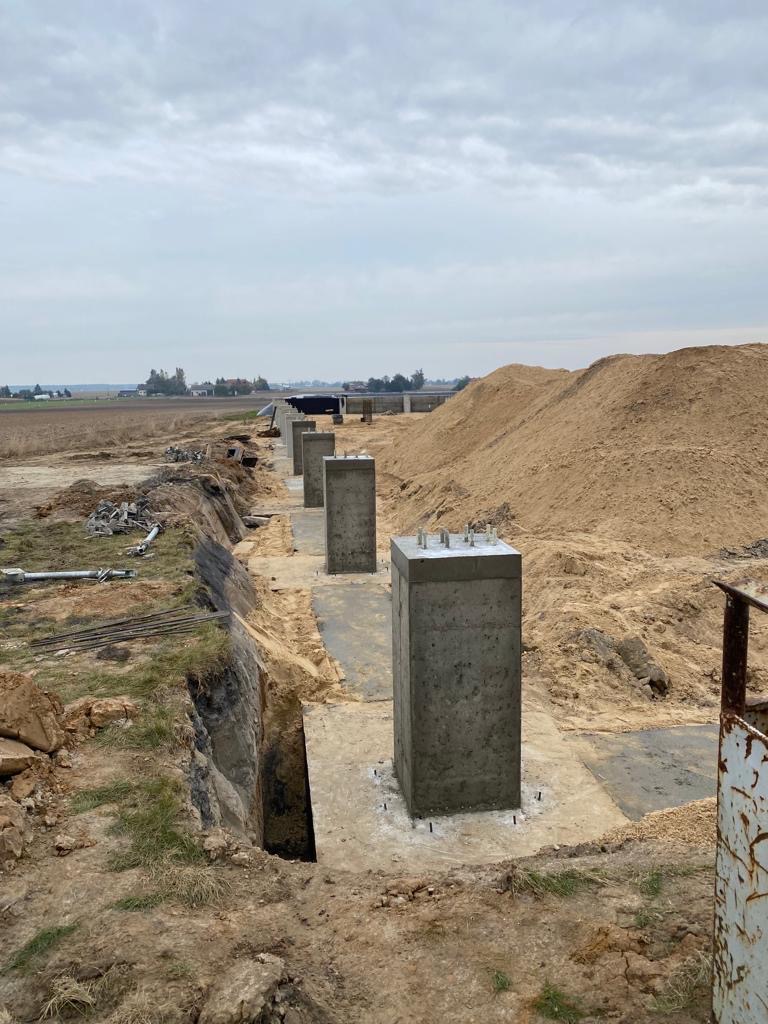 Wylane betonowe słupy fundamentowe z wystającymi prętami zbrojeniowymi, widoczne wykopy i hałda piasku, rozległy teren budowy w tle.