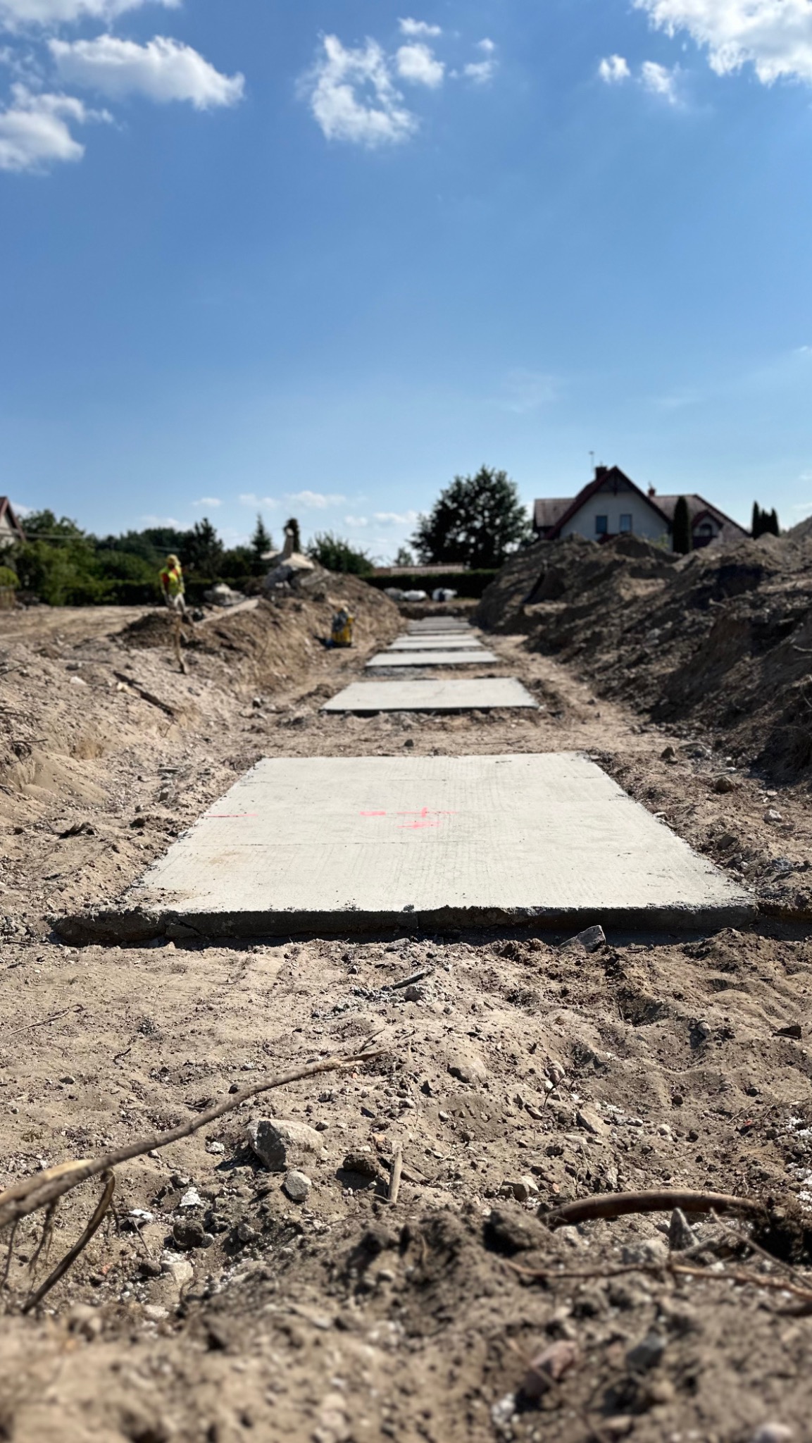 Przygotowane betonowe płyty z czerwonymi oznaczeniami ułożone w wykopie pod budowę, widoczny pracownik w oddali i błękitne niebo.