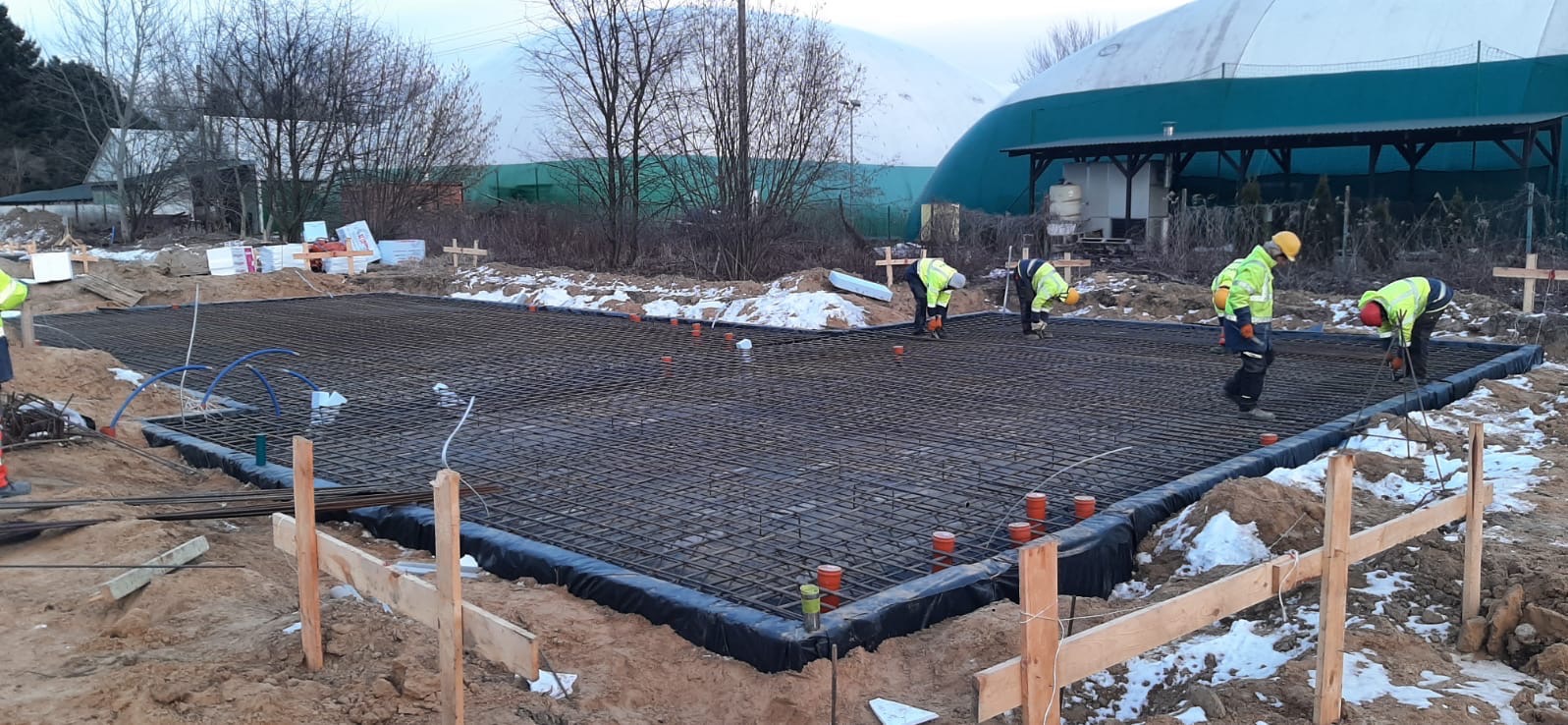 Przygotowanie zbrojenia fundamentu pod budynek; siatka z prętów stalowych, zabezpieczona folią, pomarańczowe rurki pionowe, pracownicy w odblaskowych ubraniach, w tle zima i hala sportowa.