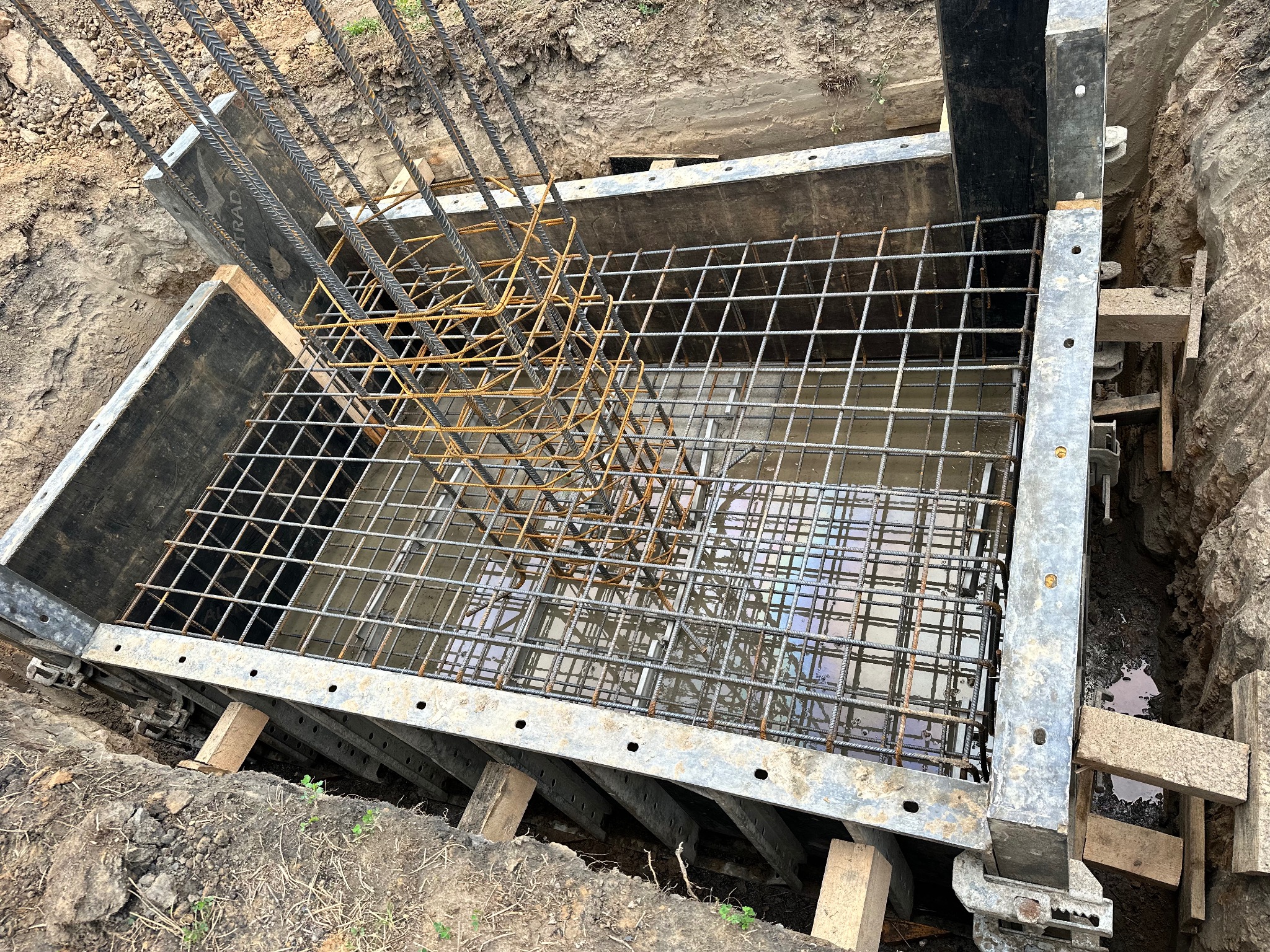 Zbrojenie fundamentu w szalunku, częściowo wypełnionym wodą, widoczne wystające pręty i siatka zbrojeniowa.