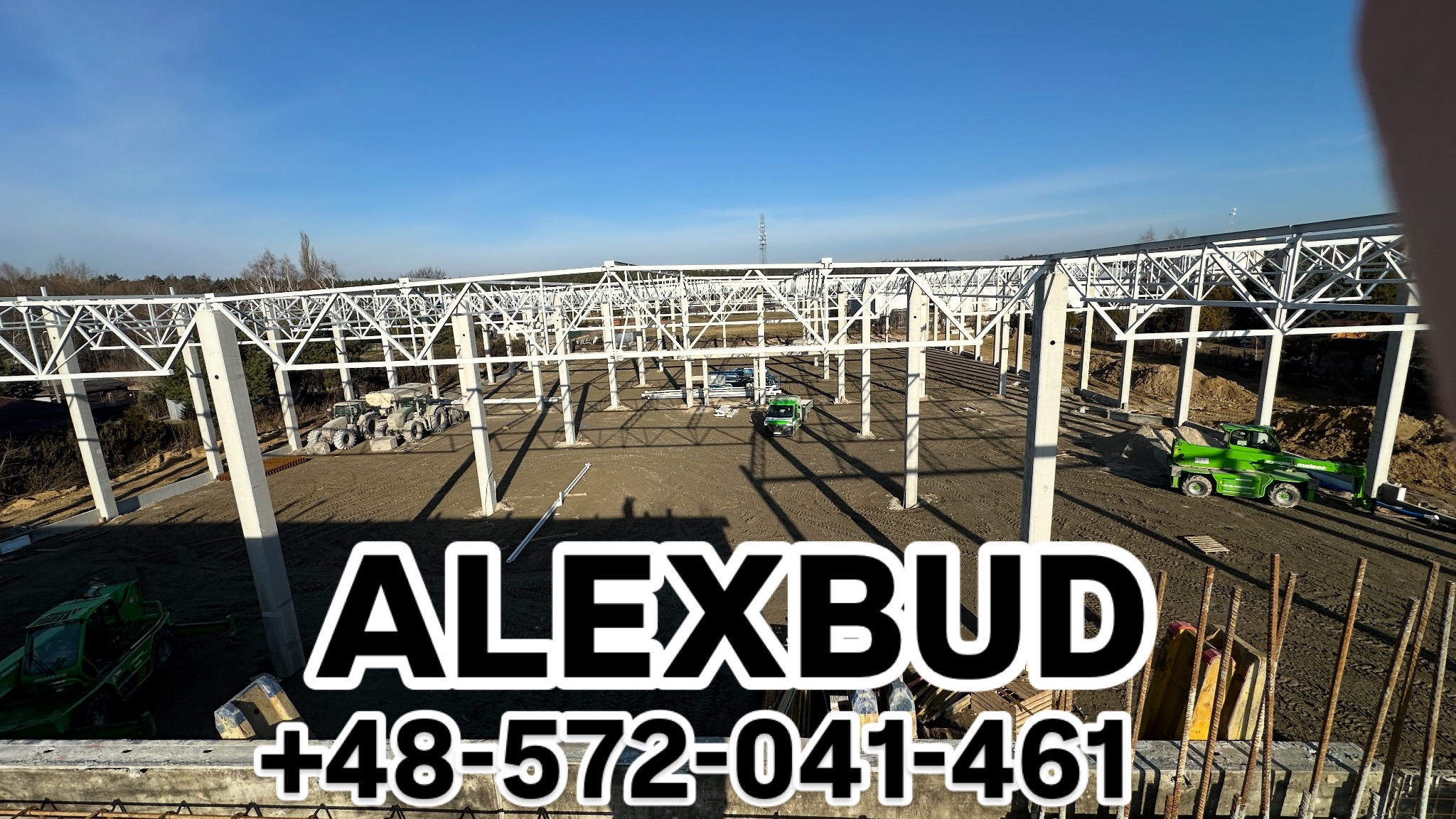 Konstrukcja stalowa hali w trakcie budowy, widoczne słupy i kratownica, sprzęt budowlany na placu budowy. Na pierwszym planie zbrojenie oraz logo firmy ALEXBUD.