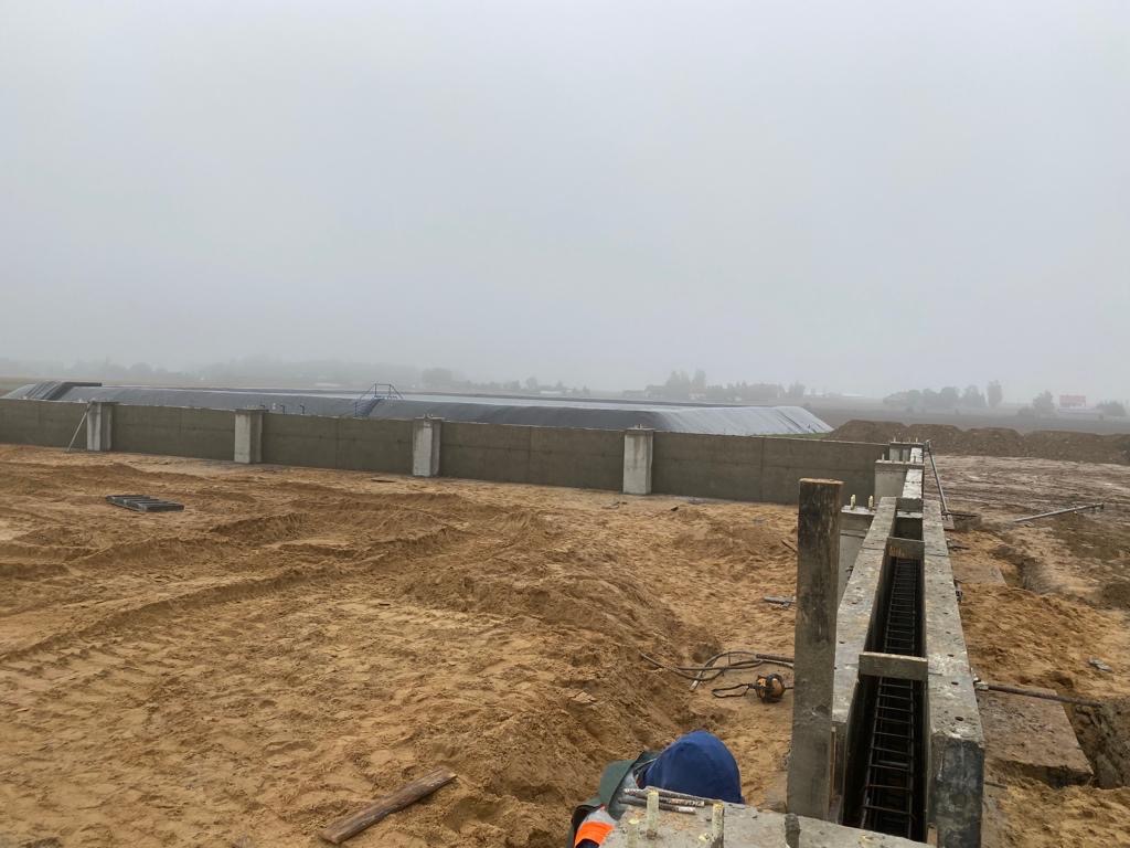 Budowa zbiornika żelbetowego w trakcie realizacji: widoczne szalunki, zbrojenia i izolacja pionowa na tle zamglonego krajobrazu.