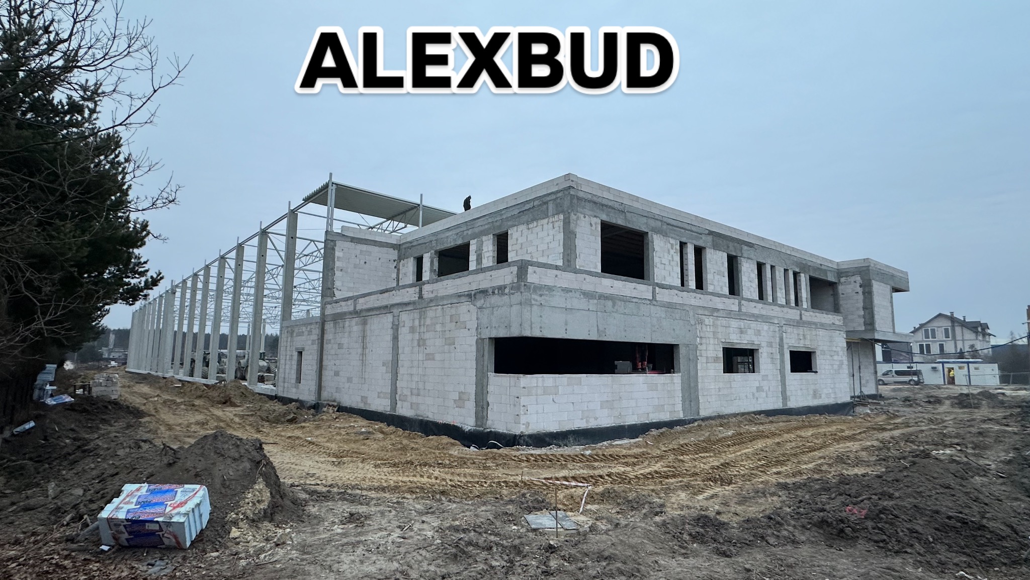 Budowa budynku z betonu komórkowego w stanie surowym otwartym, widoczne stalowe elementy konstrukcyjne oraz teren budowy z ziemią i drzewami w tle, logo firmy ALEXBUD na górze zdjęcia.