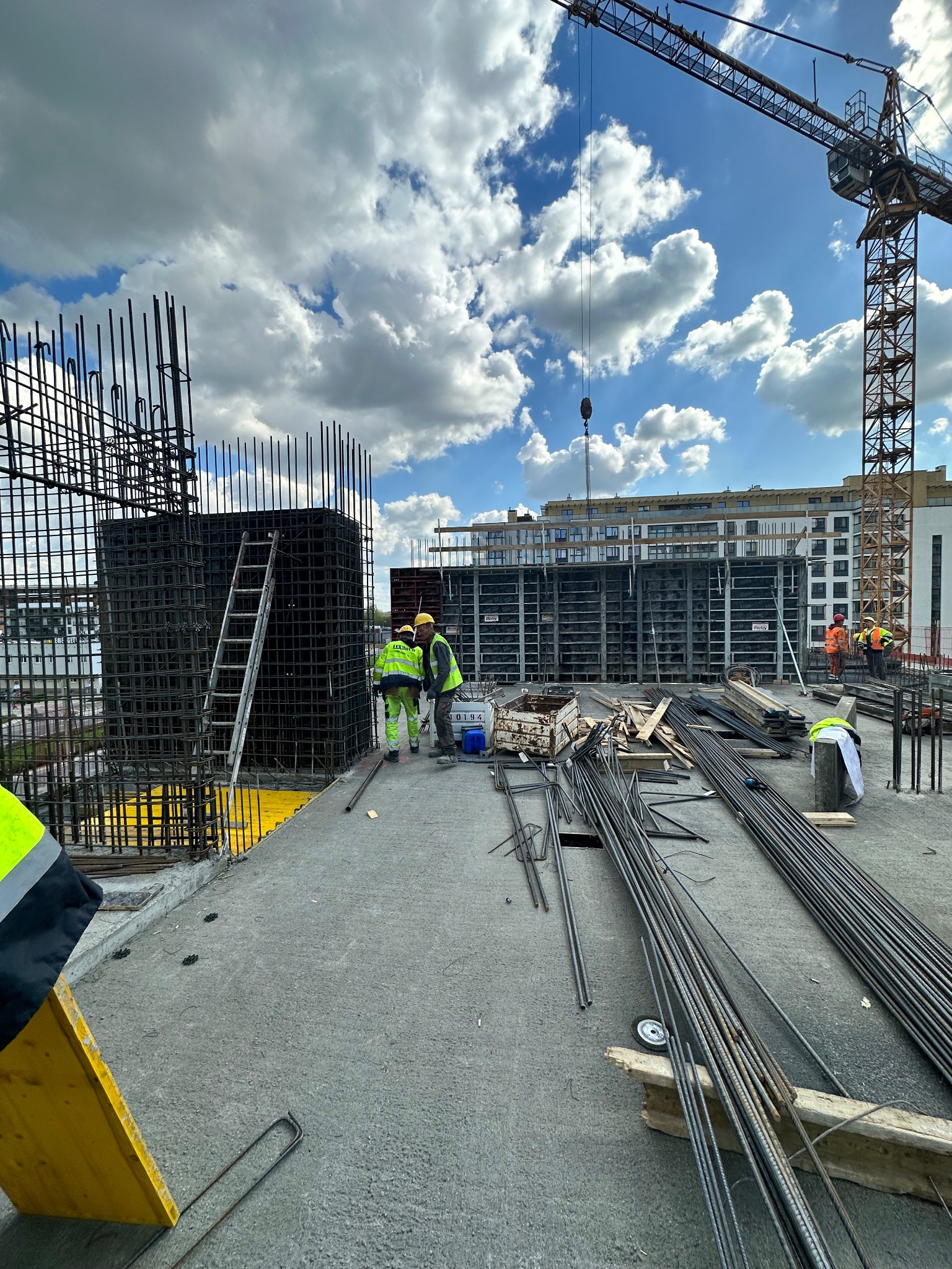 Wiązanie zbrojenia na budowie budynku wielorodzinnego, widoczne szalunki, pręty zbrojeniowe, dźwig budowlany i pracownicy w odblaskowych kamizelkach na tle błękitnego nieba z chmurami.