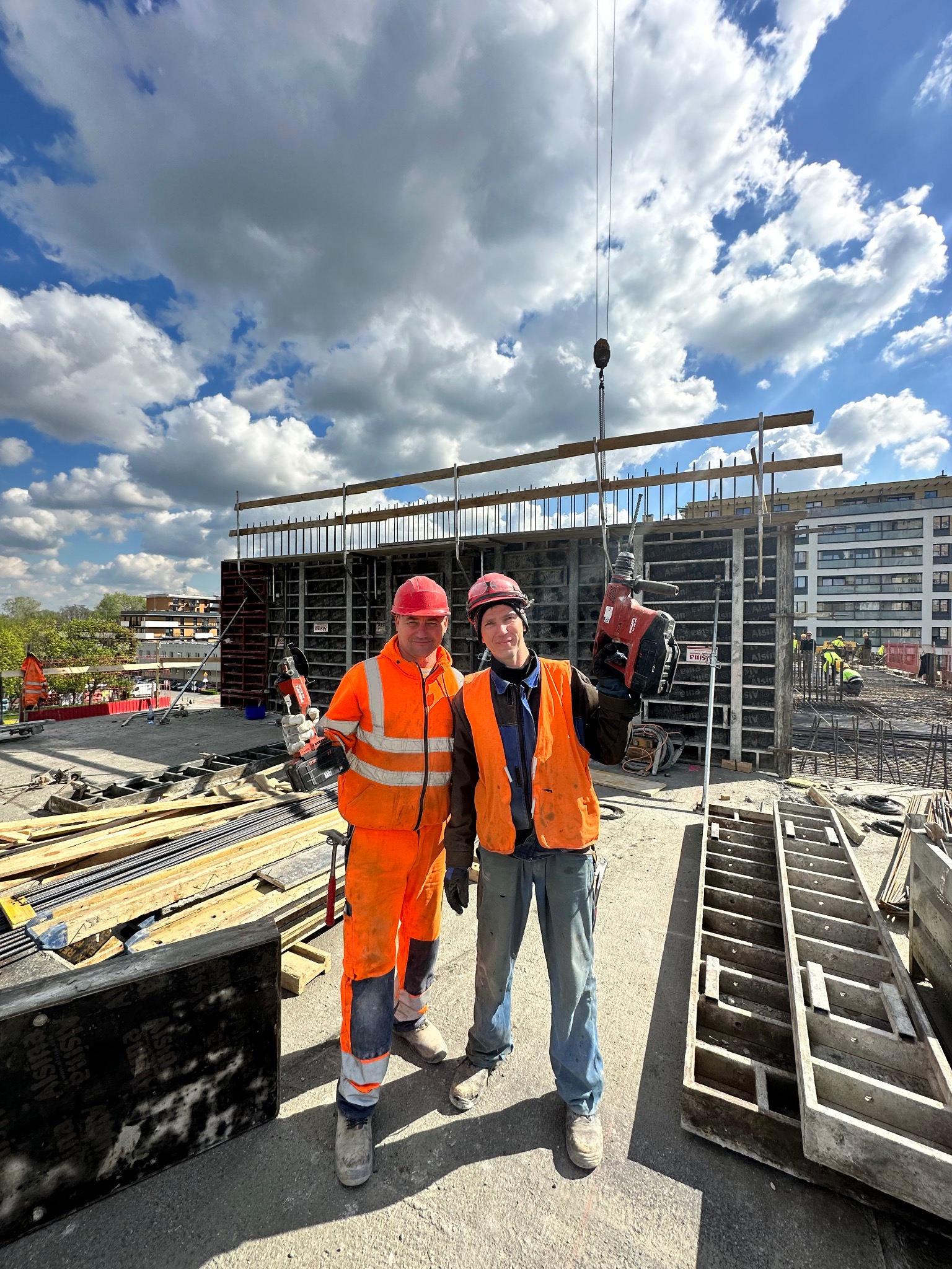 Dwóch uśmiechniętych pracowników budowlanych w pomarańczowych kombinezonach i kaskach, trzyma w rękach narzędzia do zbrojenia na tle szalunków i budowy budynku żelbetowego pod błękitnym niebem.