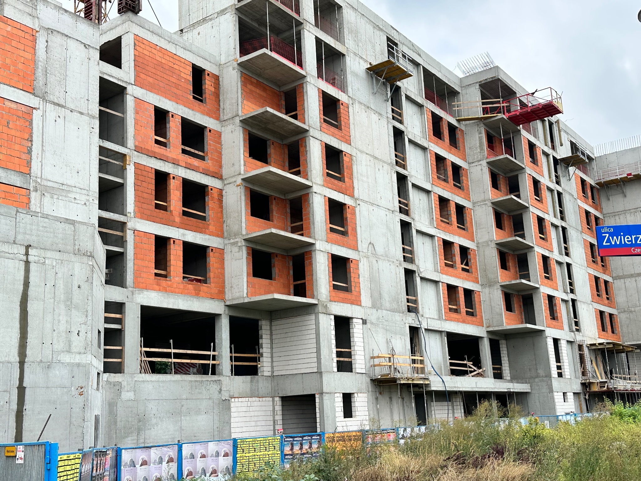 Wielopiętrowy budynek w stanie surowym, z widocznymi elementami konstrukcji żelbetowej i fragmentami muru z cegły ceramicznej, z zamontowanymi platformami roboczymi na elewacji.