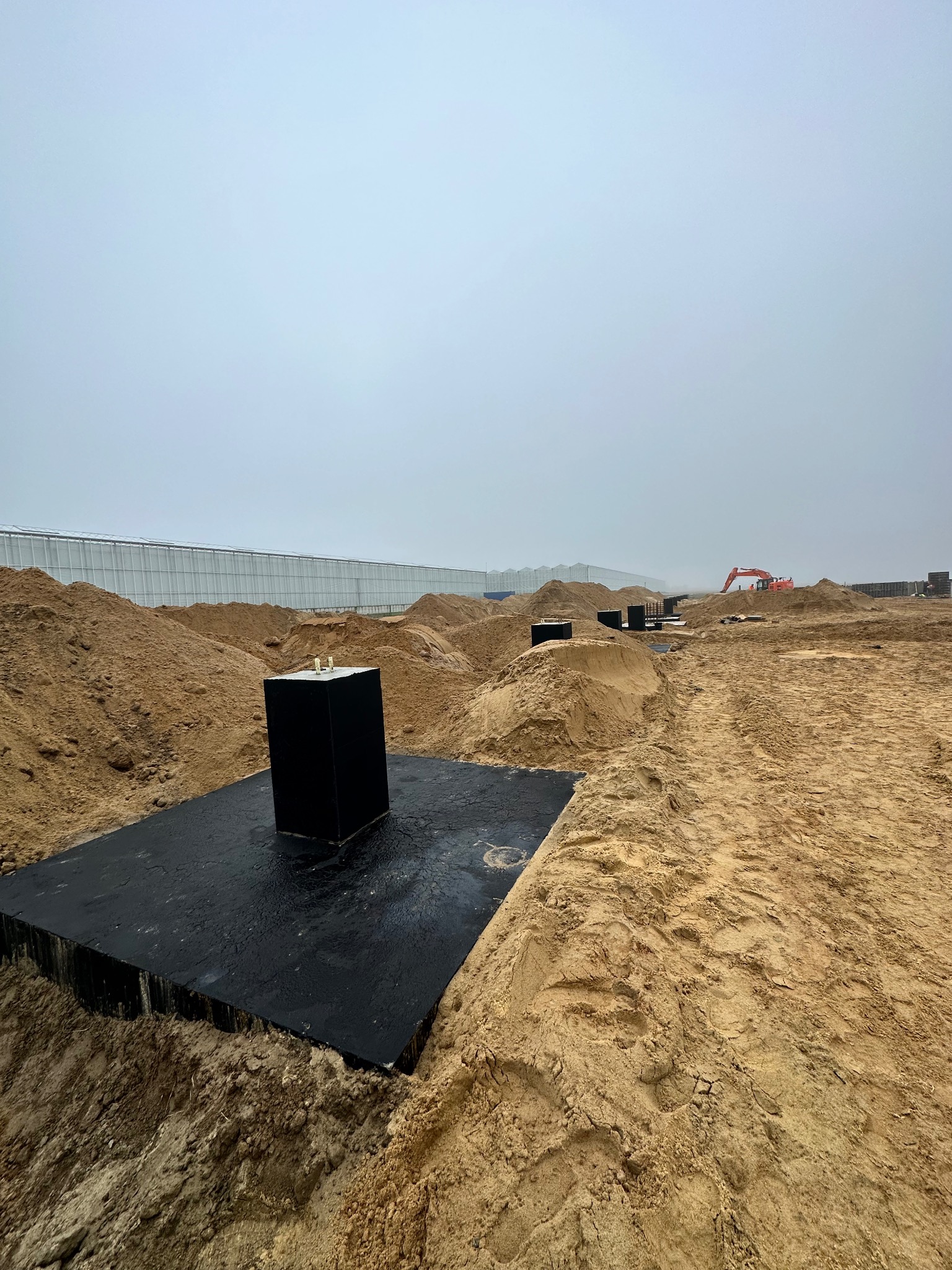 Przygotowanie terenu pod budowę, widoczne wykopane fundamenty, teren piaszczysty, w tle szklarnia i koparka.