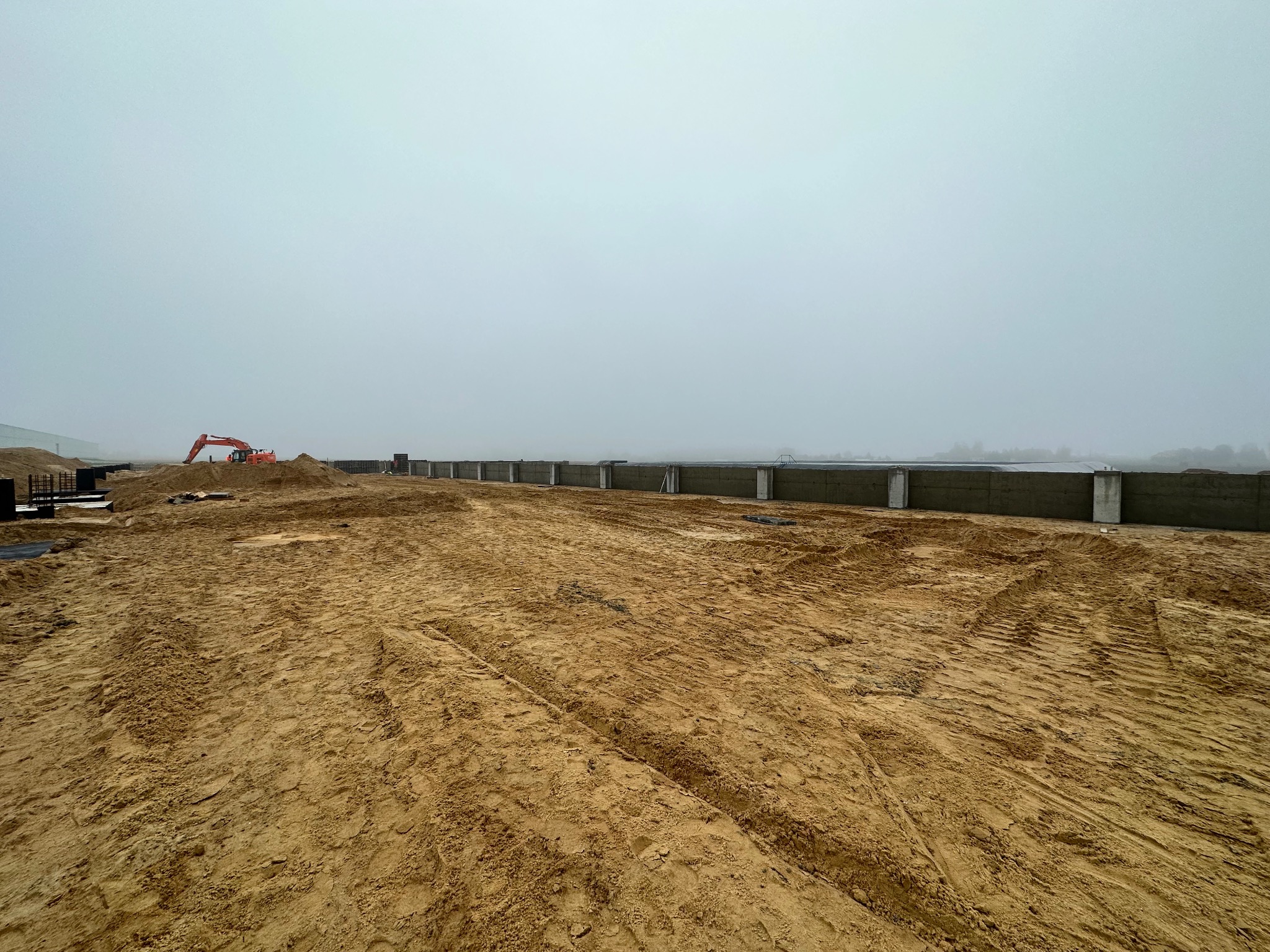 Szeroki kadr na plac budowy w mglisty dzień; widoczne przygotowane fundamenty pod budynek, wykopana ziemia z wyraźnymi śladami opon i koparka w oddali.