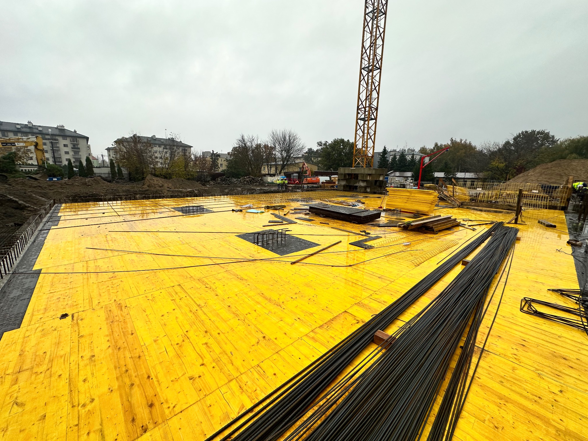 Żółte szalunki na budowie z widoczną stertą prętów zbrojeniowych i żurawiem w tle, pochmurny dzień.
