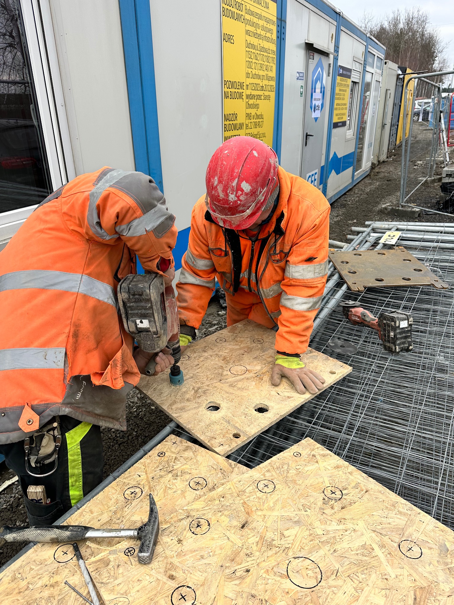 Dwóch pracowników w pomarańczowych kombinezonach wierci otwory w płytach OSB na placu budowy, widoczne narzędzia: wiertarka, młotek, gwoździe, w tle kontenery budowlane i elementy zbrojenia.