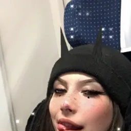 Kobieta z makijażem i czarną czapką z rogami, siedząca w pociągu, wystawia język, z efektem błyszczących gwiazdek na zdjęciu.