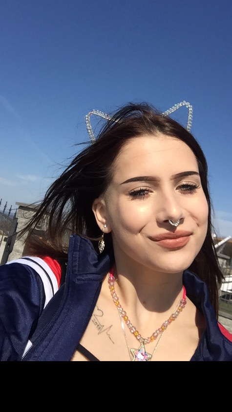 Portret uśmiechniętej brunetki z kolczykiem w nosie, w uszach i opaską z kocimi uszami na tle błękitnego nieba.