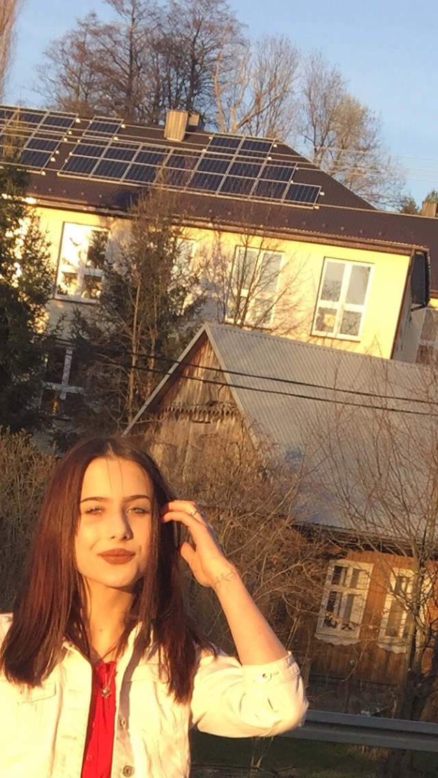 Młoda kobieta w białej kurtce na tle domu z panelami słonecznymi na dachu, w promieniach zachodzącego słońca.