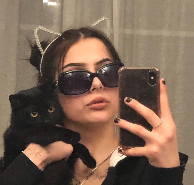 Selfie dziewczyny w okularach przeciwsłonecznych i opasce z kocimi uszami, trzymającej czarnego kota na rękach.
