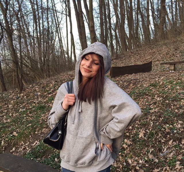 Uśmiechnięta młoda kobieta w szarej bluzie z kapturem, z czarną lakierowaną torebką na ramieniu, stoi na tle leśnego krajobrazu z opadłymi liśćmi i drzewami bez liści.