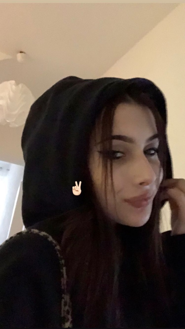 Portret kobiety w czarnej bluzie z kapturem, z delikatnym makijażem oczu i ust, uśmiechnięta, z dłonią przy twarzy