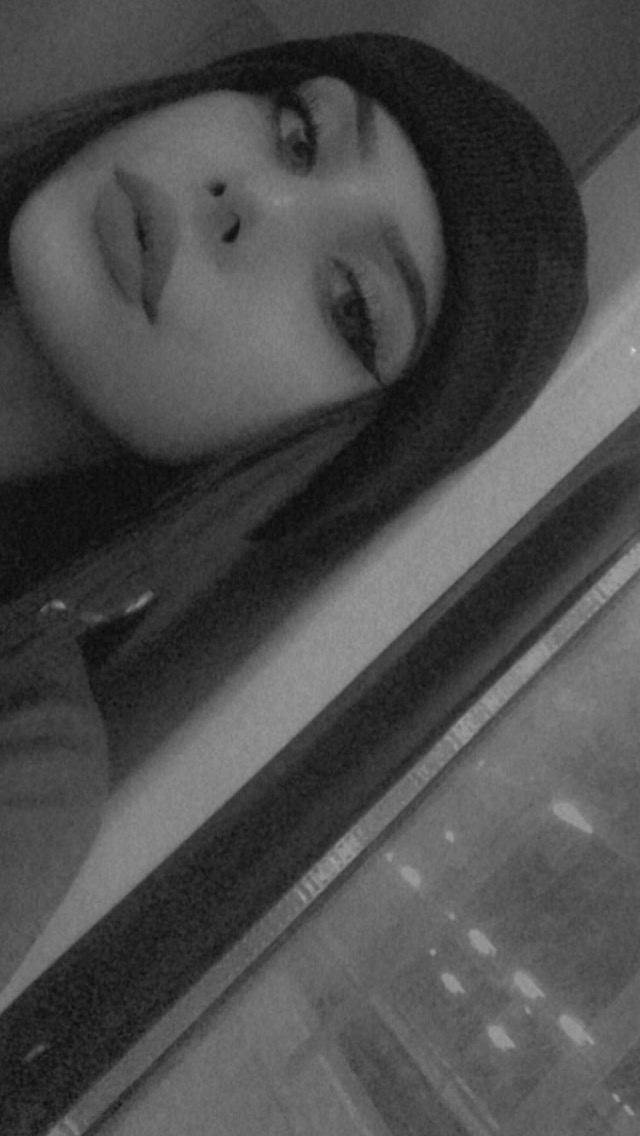 Portret kobiety w czarno-białej tonacji, z wyraźnie podkreślonym makijażem oczu i ustami skierowanymi w stronę obiektywu, w tle rozmyte światła miasta za oknem.