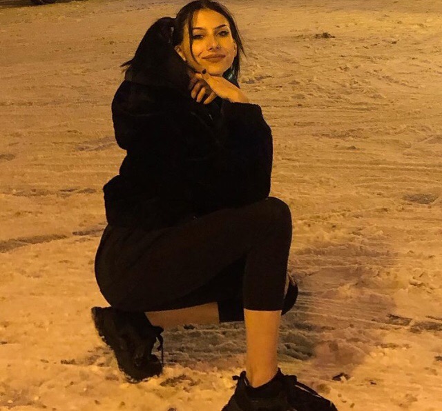 Kobieta w czarnym dresie kuca na śniegu, opierając brodę na dłoniach i uśmiechając się w kierunku kamery.