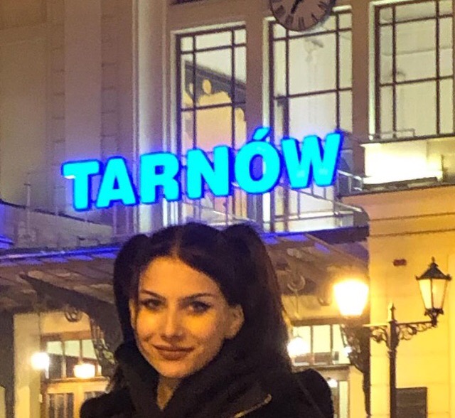 Portret uśmiechniętej kobiety na tle budynku dworca kolejowego z neonowym napisem 'Tarnów'.