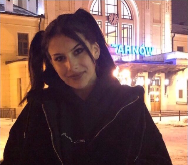 Portret młodej kobiety z ciemnymi włosami upiętymi w dwa kucyki, z wyraźnym makijażem oczu, na tle oświetlonego dworca kolejowego w Tarnowie nocą.