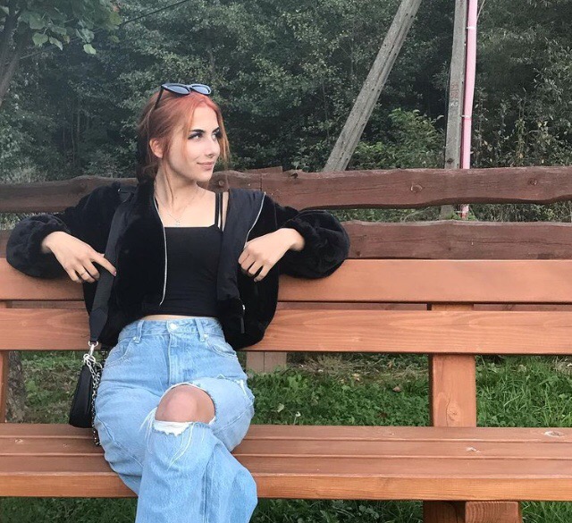 Kobieta z rudymi włosami i okularami na głowie siedzi na drewnianej ławce w parku, ubrana w czarną kurtkę i podarte jeansy.