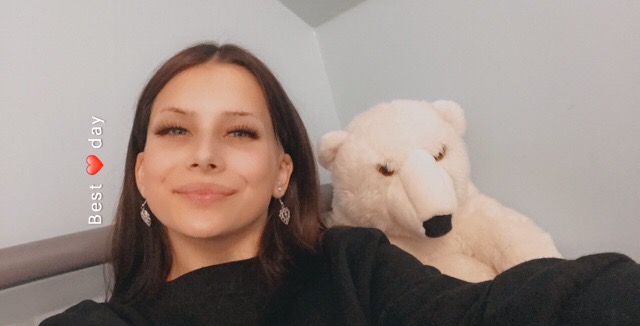 Uśmiechnięta brunetka z długimi rzęsami i kolczykami w kształcie liści, pozująca z pluszowym białym misiem polarnym na tle jasnej ściany.