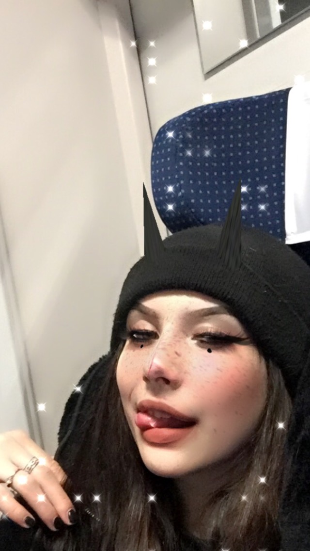 Kobieta z makijażem i czarną czapką z rogami, siedząca w pociągu, wystawia język, z efektem błyszczących gwiazdek na zdjęciu.