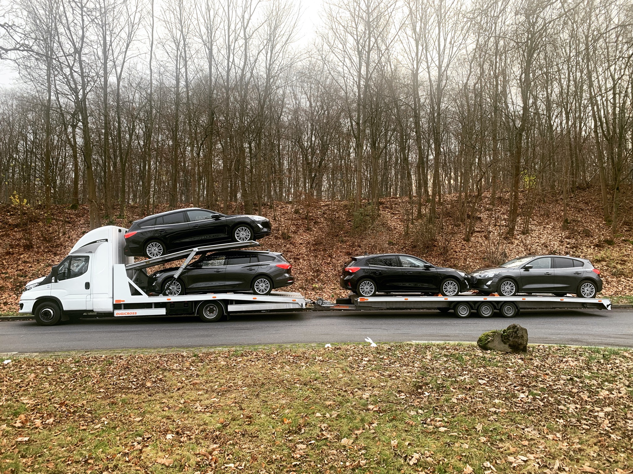 Biała laweta transportuje cztery ciemne samochody osobowe na dwóch poziomach, tło stanowią drzewa bez liści i opadłe liście.