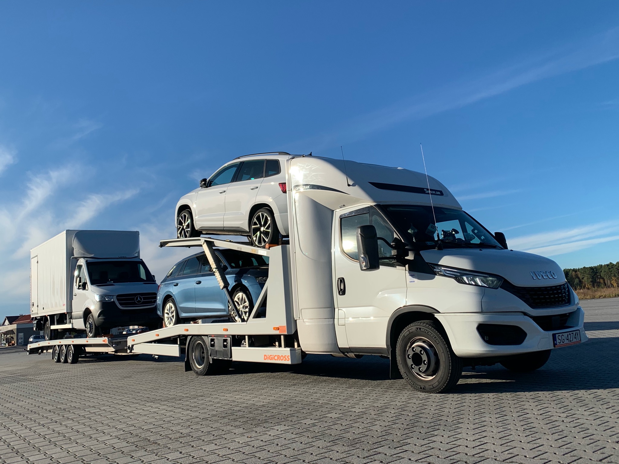 Laweta z dwoma samochodami osobowymi, biały SUV na górze i niebieskie kombi na dole, ciągnięta przez biały samochód ciężarowy marki Iveco, na pierwszym planie brukowa nawierzchnia, w tle błękitne...