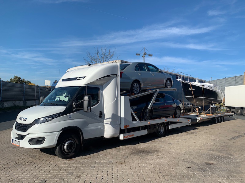 Biała laweta Iveco z dwoma samochodami osobowymi i czarną łodzią na platformie, zaparkowana na brukowanym placu w słoneczny dzień.