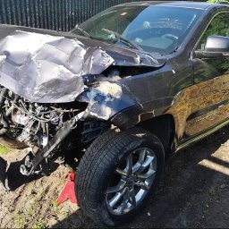 Dzinks - Uszkodzony przód czarnego Jeepa Grand Cherokee po kolizji, widoczne zniszczenia karoserii i odsłonięte elementy silnika, auto stoi na ziemi.