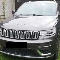 Dzinks - Szary Jeep Grand Cherokee z chromowanym grillem, zaparkowany na trawie przed garażem. Widok z przodu, lekko z boku. Czysty, zadbany wygląd.