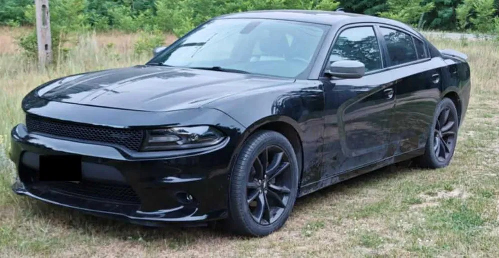 Czarny samochód osobowy, Dodge Charger, zaparkowany na trawie. Widok z przodu i boku, zarys lasu w tle. Auto po detailingu, z połyskiem lakieru.