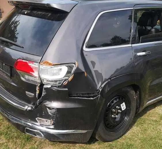 Uszkodzony, ciemny SUV po kolizji: wgniecione blachy, zarysowany lakier, pęknięty reflektor. Widoczne ślady rdzy. Koło zapasowe. Trawa w tle.