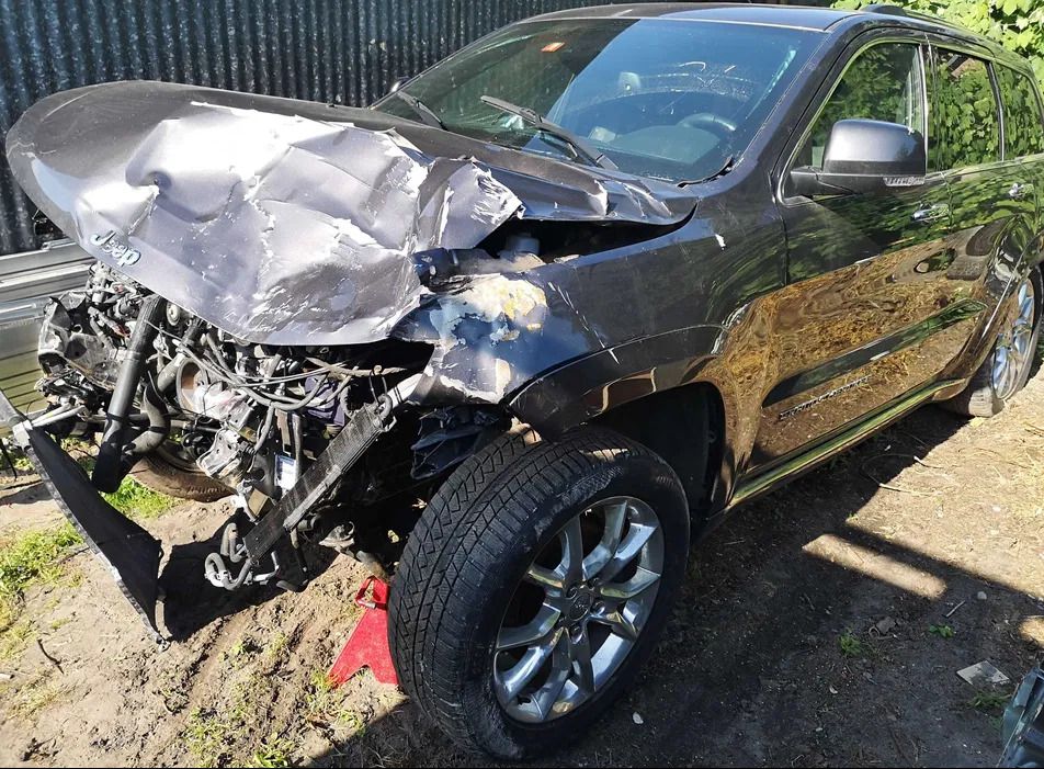 Uszkodzony przód czarnego Jeepa Grand Cherokee po kolizji, widoczne zniszczenia karoserii i odsłonięte elementy silnika, auto stoi na ziemi.