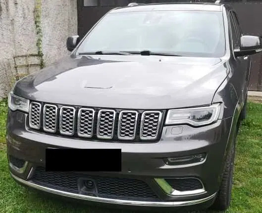 Szary Jeep Grand Cherokee z chromowanym grillem, zaparkowany na trawie przed garażem. Widok z przodu, lekko z boku. Czysty, zadbany wygląd.