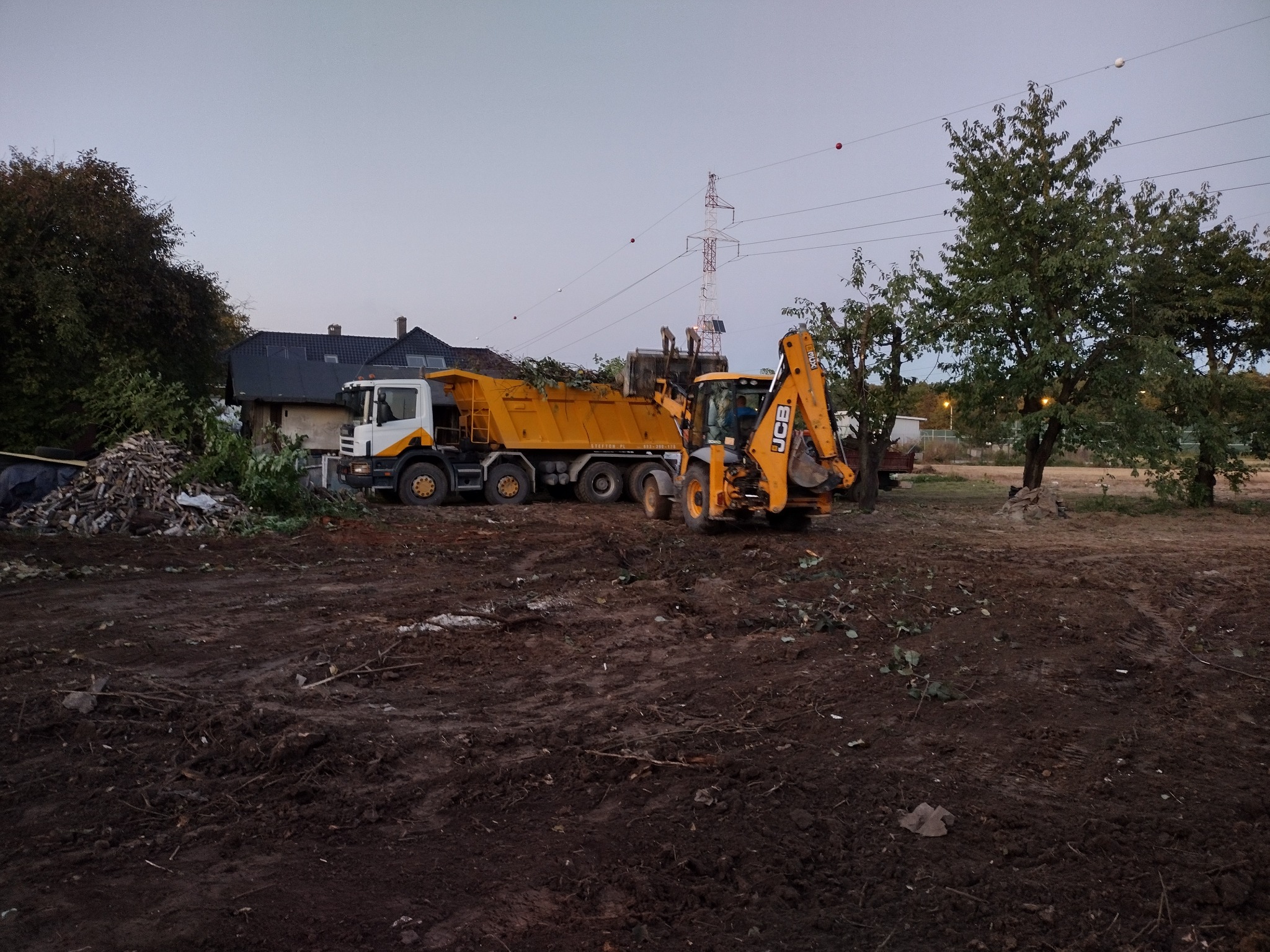 Żółta wywrotka i koparko-ładowarka JCB na placu budowy, w tle sterta pociętych gałęzi i fragment budynku.