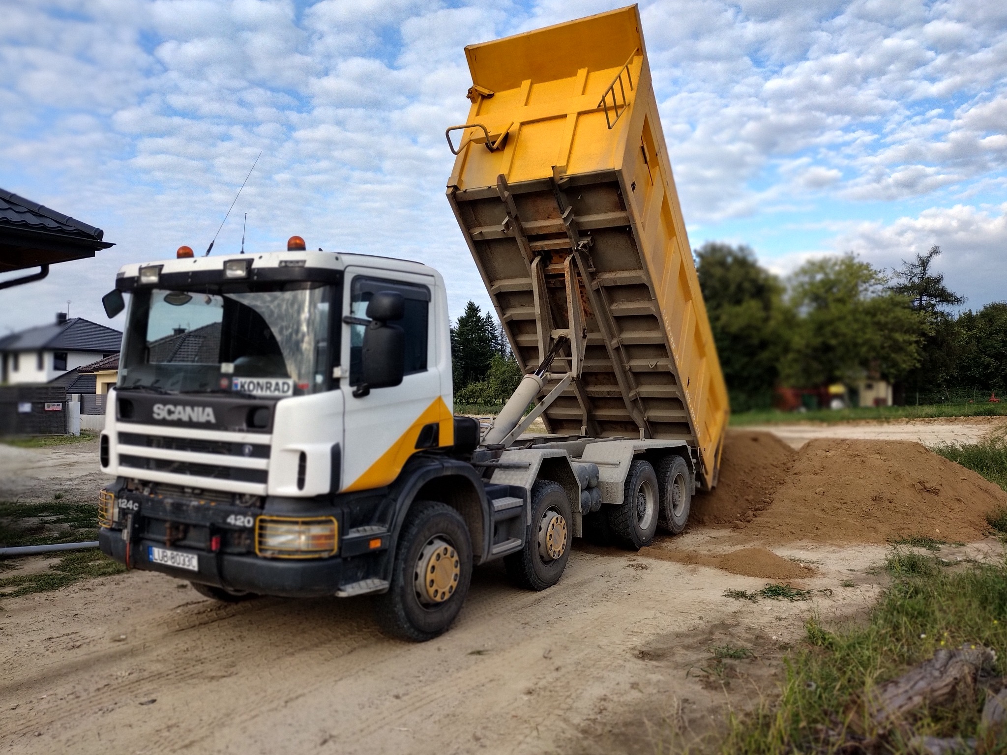Żółta wywrotka Scania 420 z podniesioną skrzynią ładunkową wysypuje brązowy materiał sypki na placu budowy, widoczny numer rejestracyjny LUB-8033C.