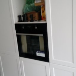 marek witkowski - Biała zabudowa kuchenna z piekarnikiem w trakcie montażu, widoczne kartony i akcesoria na górnej półce.