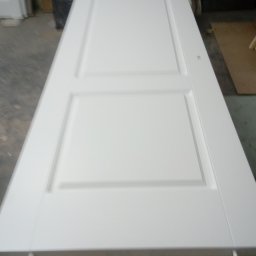 marek witkowski - Białe, dwupanelowe drzwi wewnętrzne leżące poziomo na drewnianym stojaku w warsztacie, widoczne ślady obróbki.