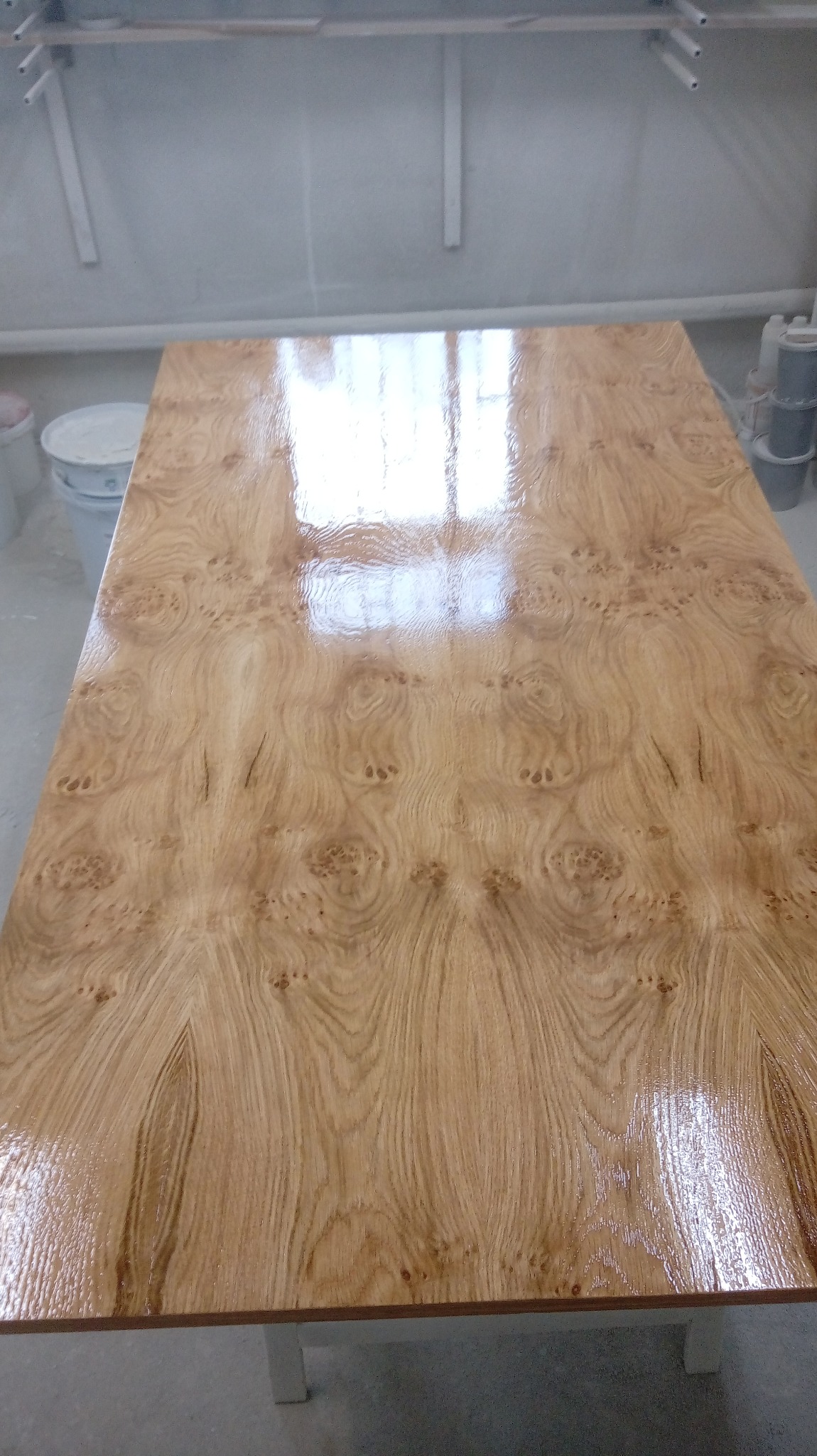 Błyszczący, lakierowany blat drewniany z wyraźnym usłojeniem, odbijający światło okna w warsztacie.