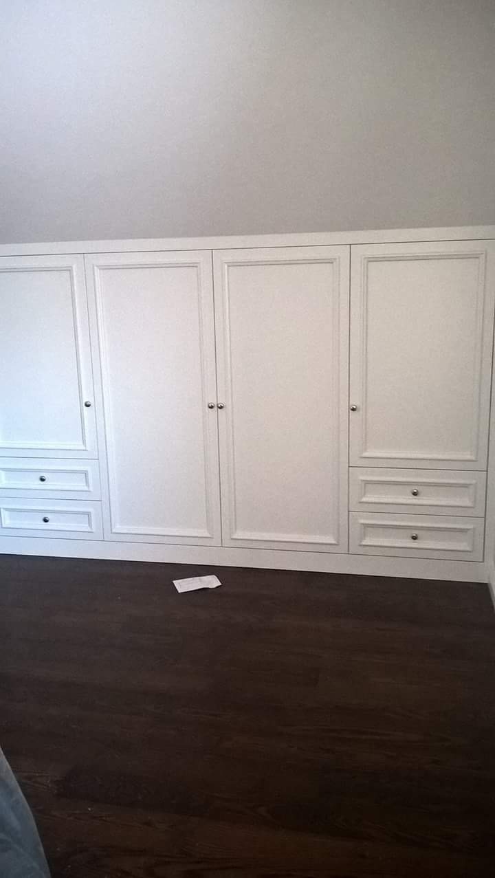 Biała szafa wnękowa z szufladami i srebrnymi gałkami, widoczna na tle ciemnej podłogi, z niedbale rzuconą kartką na podłodze.