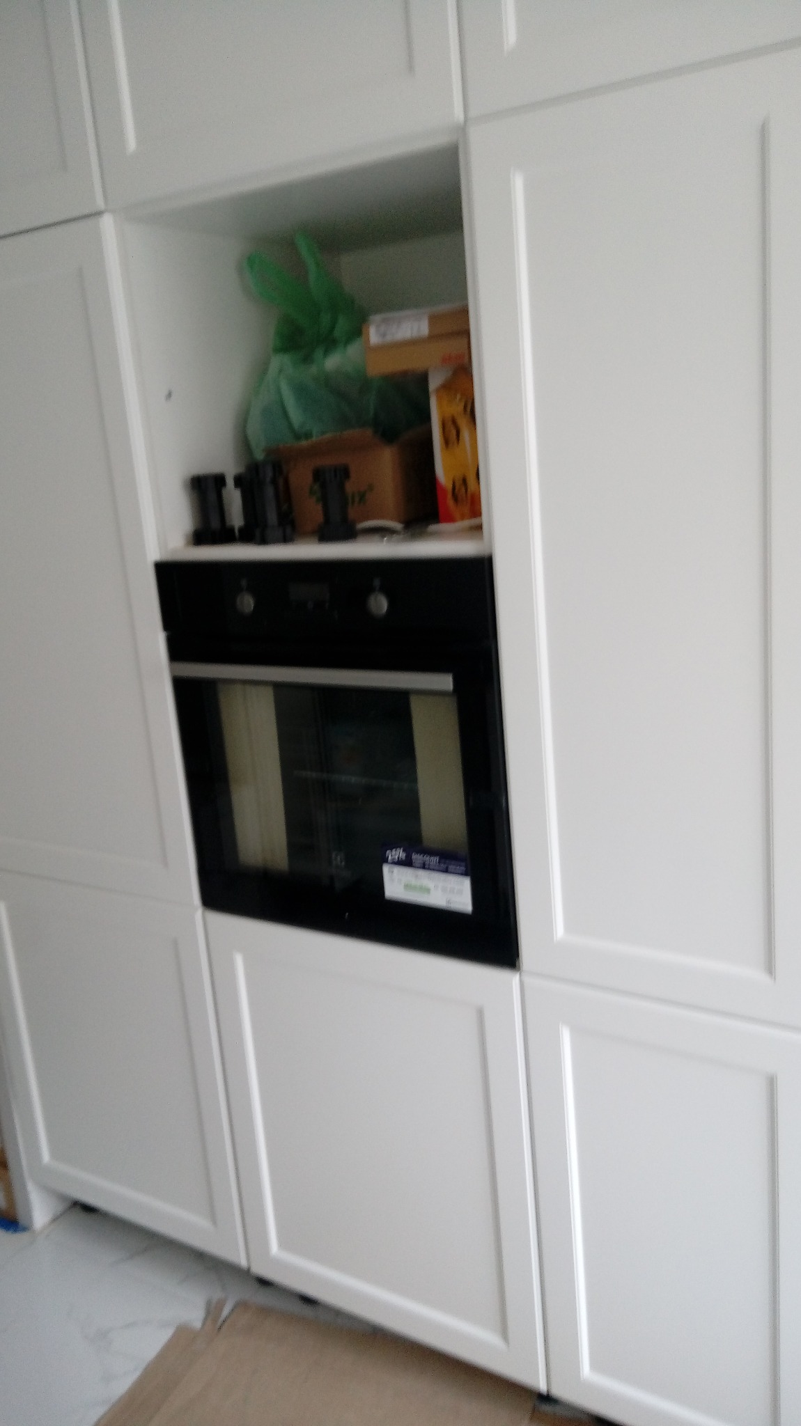 Biała zabudowa kuchenna z piekarnikiem w trakcie montażu, widoczne kartony i akcesoria na górnej półce.