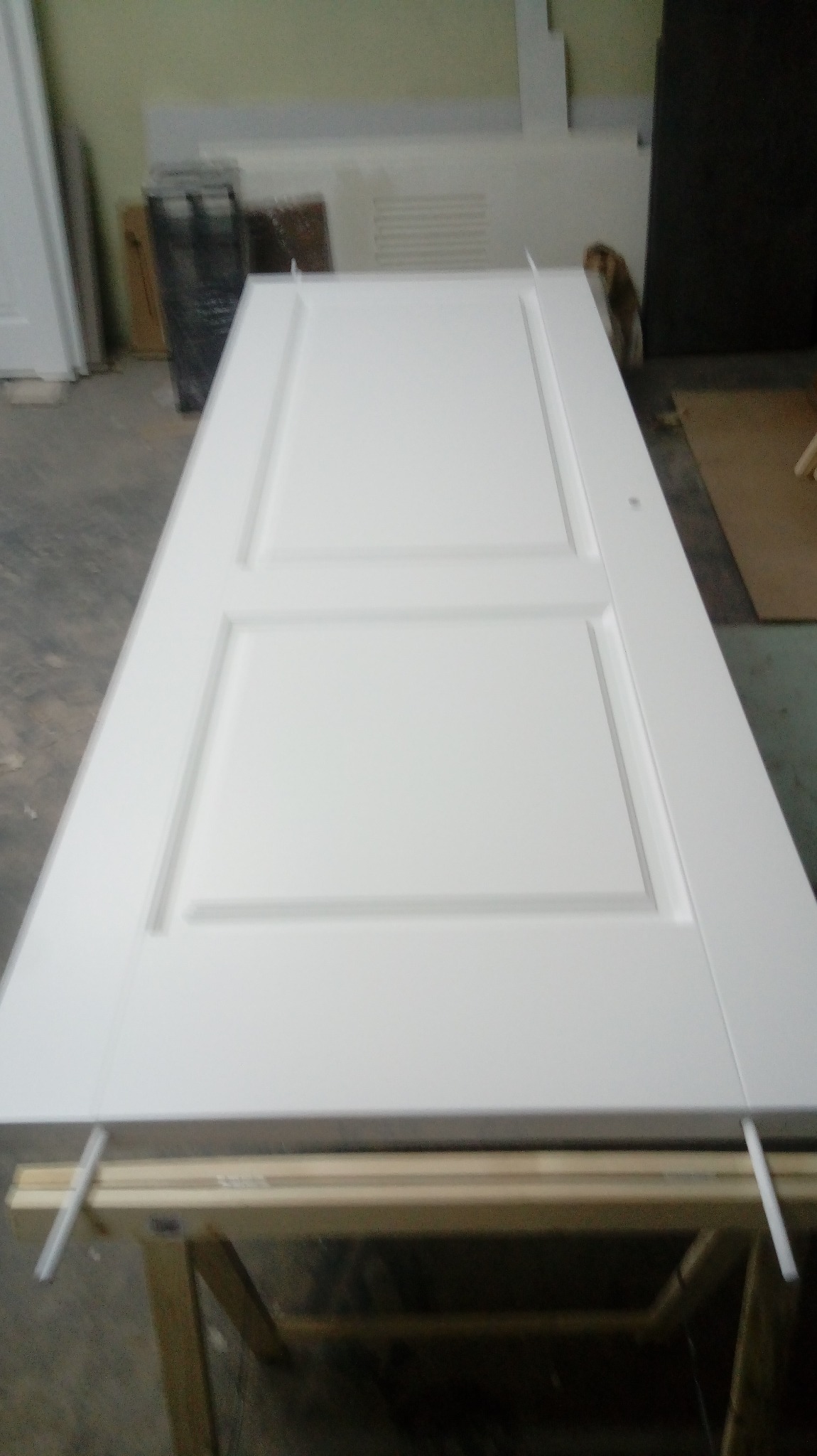 Białe, dwupanelowe drzwi wewnętrzne leżące poziomo na drewnianym stojaku w warsztacie, widoczne ślady obróbki.