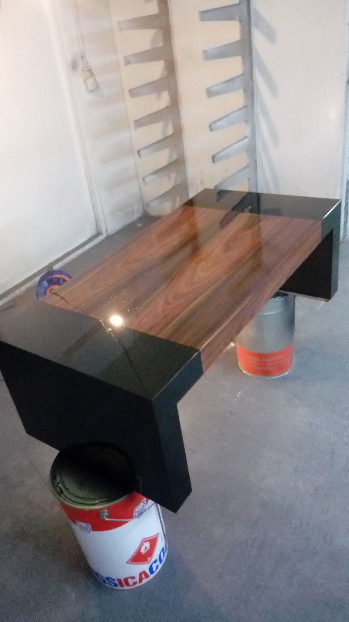 Blat drewniany z czarnymi, lakierowanymi nogami, ustawiony na puszkach farby w warsztacie, ukazujący proces wykończeniowy.