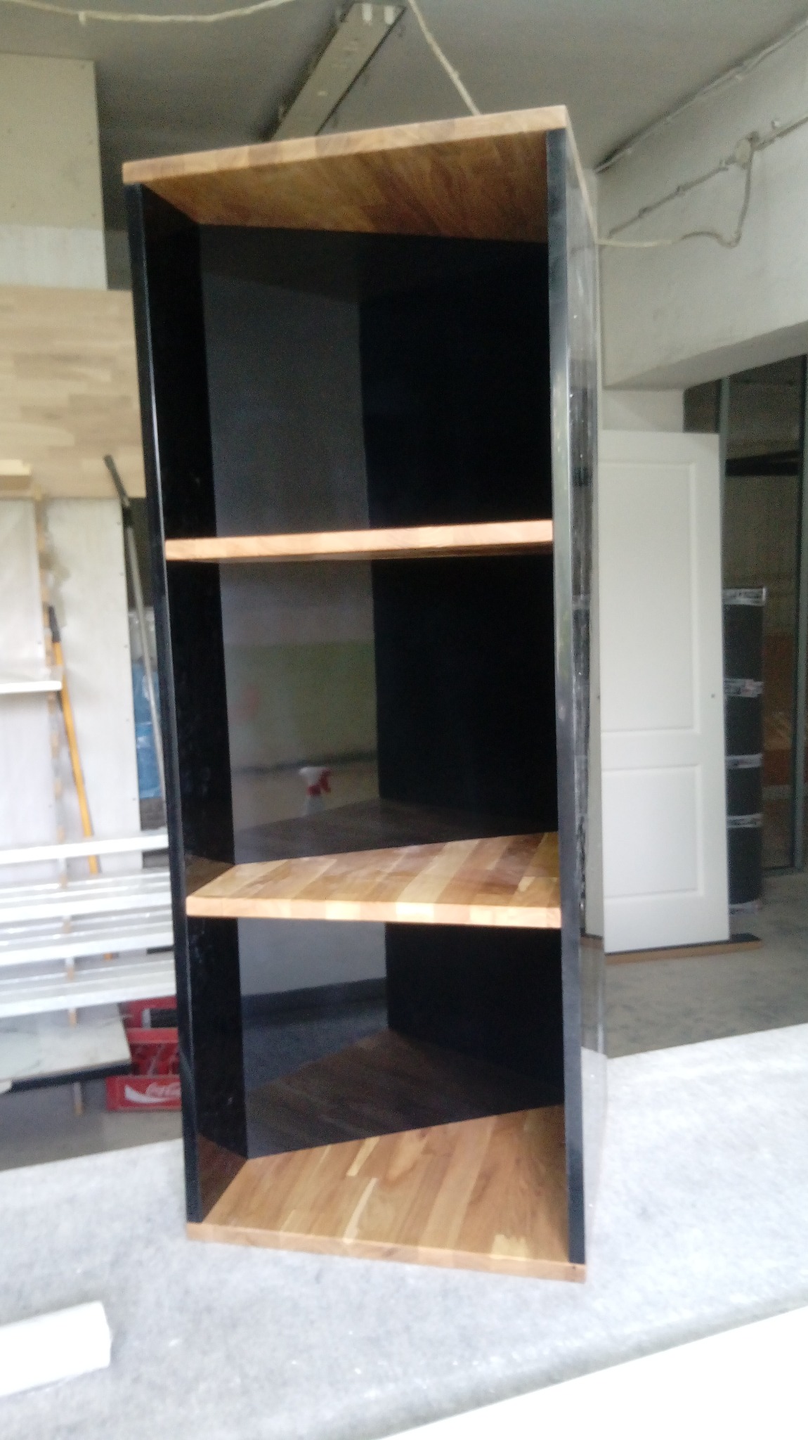 Częściowo zmontowana, czarno-drewniana szafka z trzema półkami w warsztacie, widoczna biała ściana i drzwi w tle.