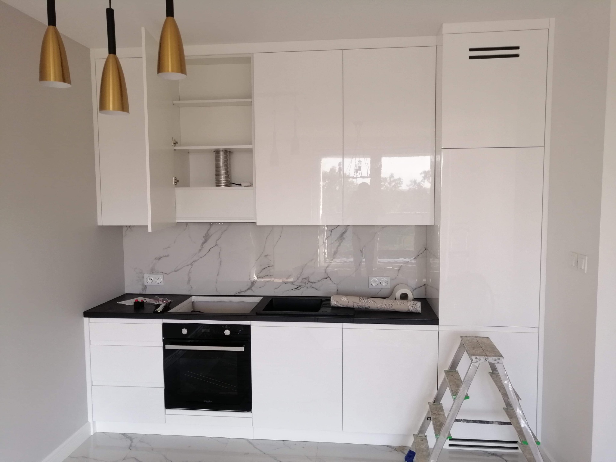 Nowoczesna, biała kuchnia z czarnym blatem i marmurowym backsplash, złote lampy wiszące, drabina w rogu wskazuje na trwający montaż.