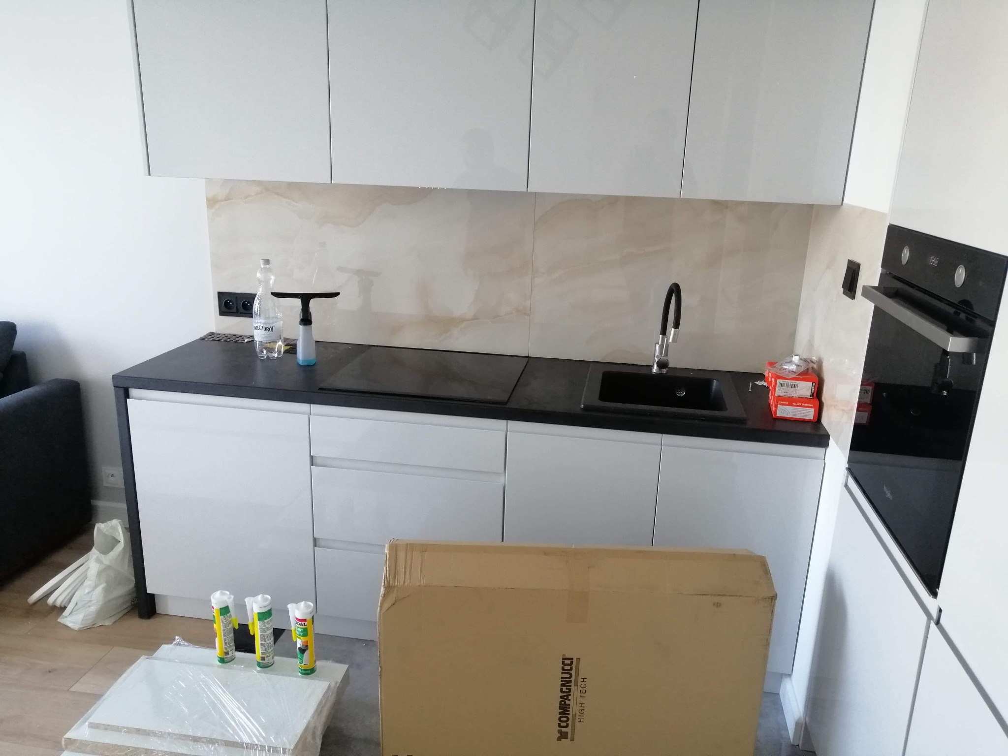 Montaż białej, nowoczesnej kuchni: widoczne szafki wiszące, ciemny blat, płytki imitujące marmur, częściowo rozpakowane elementy i tuby silikonu.