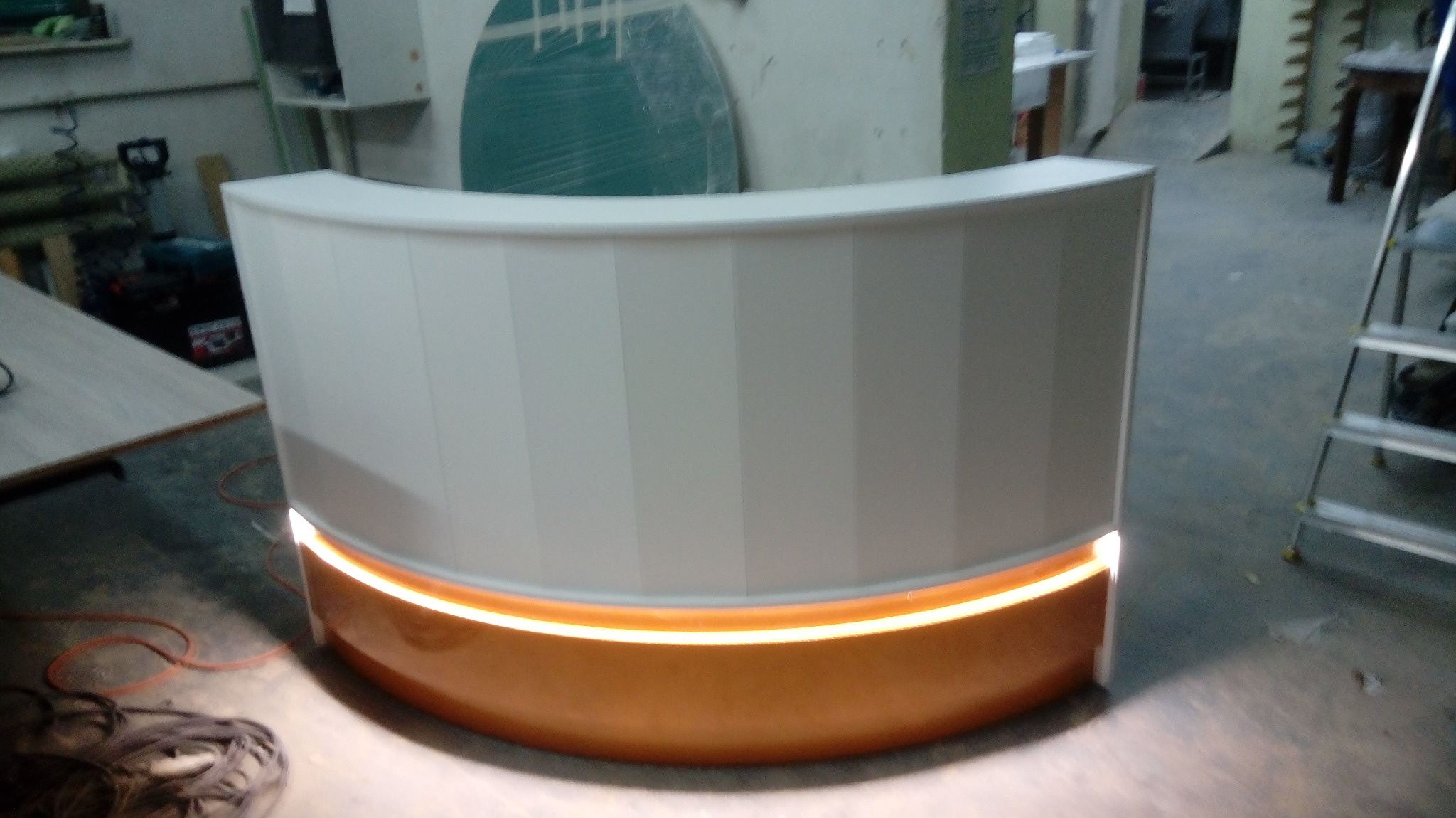 Zaokrąglony, biały front lady barowej z podświetleniem LED w kolorze ciepłego brązu, widoczne kable na podłodze i drabina w tle.